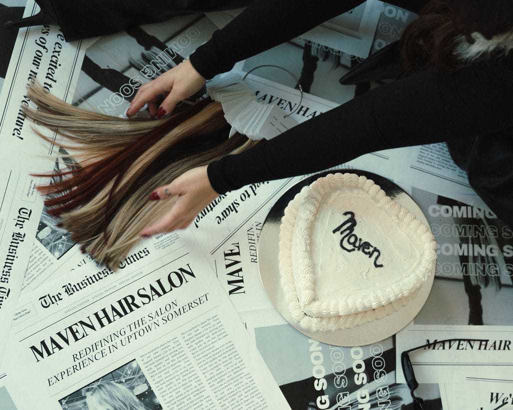 Hands holding hair color samples next to a heart-shaped cake and salon newspapers.