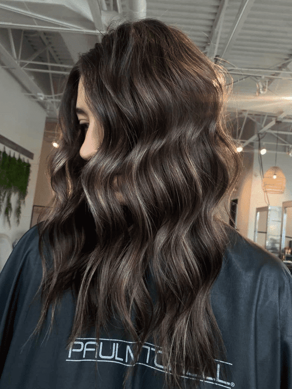 Woman with wavy, dark brown hair in a salon, wearing a black cape with "Paul Mitchell" logo.